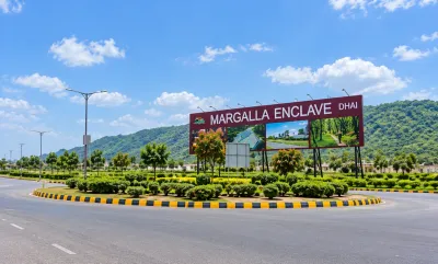 Margalla Enclave DHA Islamabad entrance sign beside a landscaped roundabout, wide road, green hills, and clear blue sky.