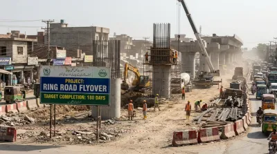 A daytime documentary photograph of heavy machinery and construction workers actively building a large concrete flyover in Peshawar, with a project sign visible.
