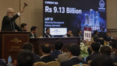 A professional documentary-style photo of the Jinnah Convention Centre interior during an auction, featuring a large digital display showing 'Rs 9.13 Billion' and 'Lot 12', with an auctioneer's gavel raised in the foreground and a focused audience in business attire.