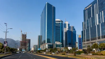 A high-end architectural photograph of the Blue Area commercial district in Islamabad, showcasing sleek glass skyscrapers, wide clean roads with light traffic, and lush greenery under a clear blue sky.