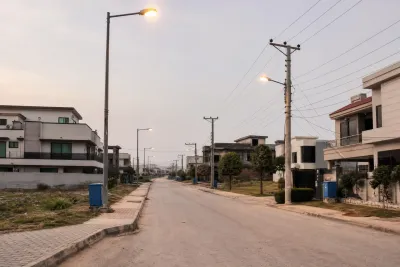 Residential street with paved road and electricity infrastructure, flanked by a mix of built and empty plots.