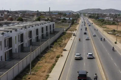 Wide road running past a housing scheme boundary wall with older industrial town rooftops in the distance.