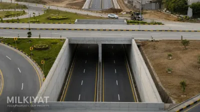 Aerial daytime view of a modern road underpass with divided lanes, landscaped medians, curved ramps, streetlights, and surrounding construction work.