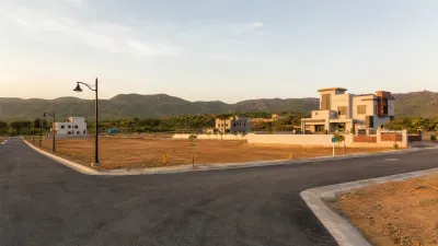 Modern houses and open residential plots along newly developed roads with green hills in the background.