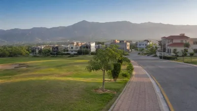 Landscaped private housing community with broad roads, manicured green space, detached homes and Margalla hills in the background.