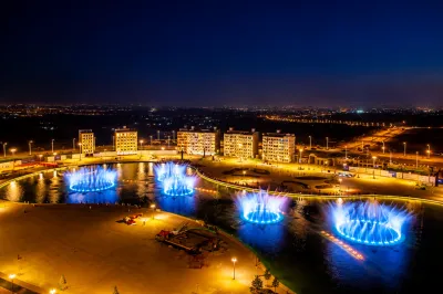 Night view of a landscaped waterfront development with illuminated buildings, roads, and blue-lit fountains.