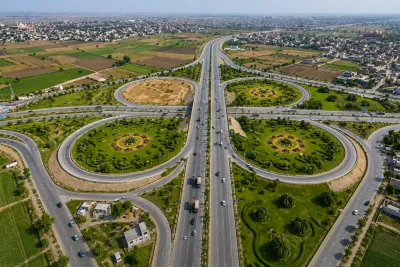 Aerial view of a major Rawalpindi Ring Road interchange with multiple looping ramps, landscaped green areas, surrounding farmland, urban development, and moving traffic.