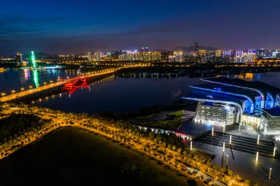Aerial night view of a modern waterfront city featuring an illuminated red bridge, high-rise buildings and a large blue-lit convention centre.