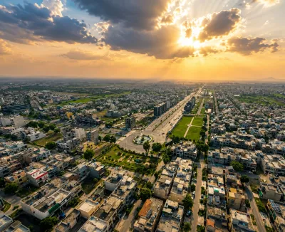 Aerial view of a developed urban area with dense housing, wide roads, landscaped green spaces, and dramatic sunset clouds.
